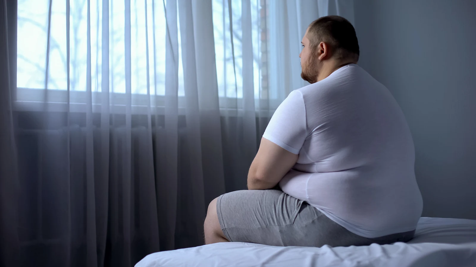 Obese man sitting on bed looking out window