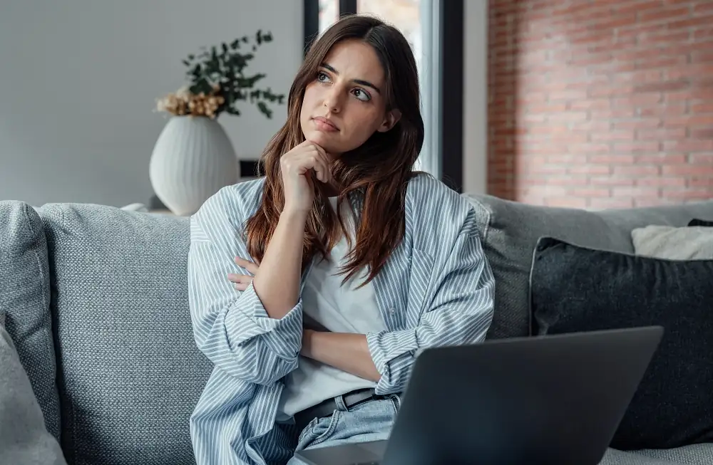 worried young woman sitting on couch with laptop, thinking of problems